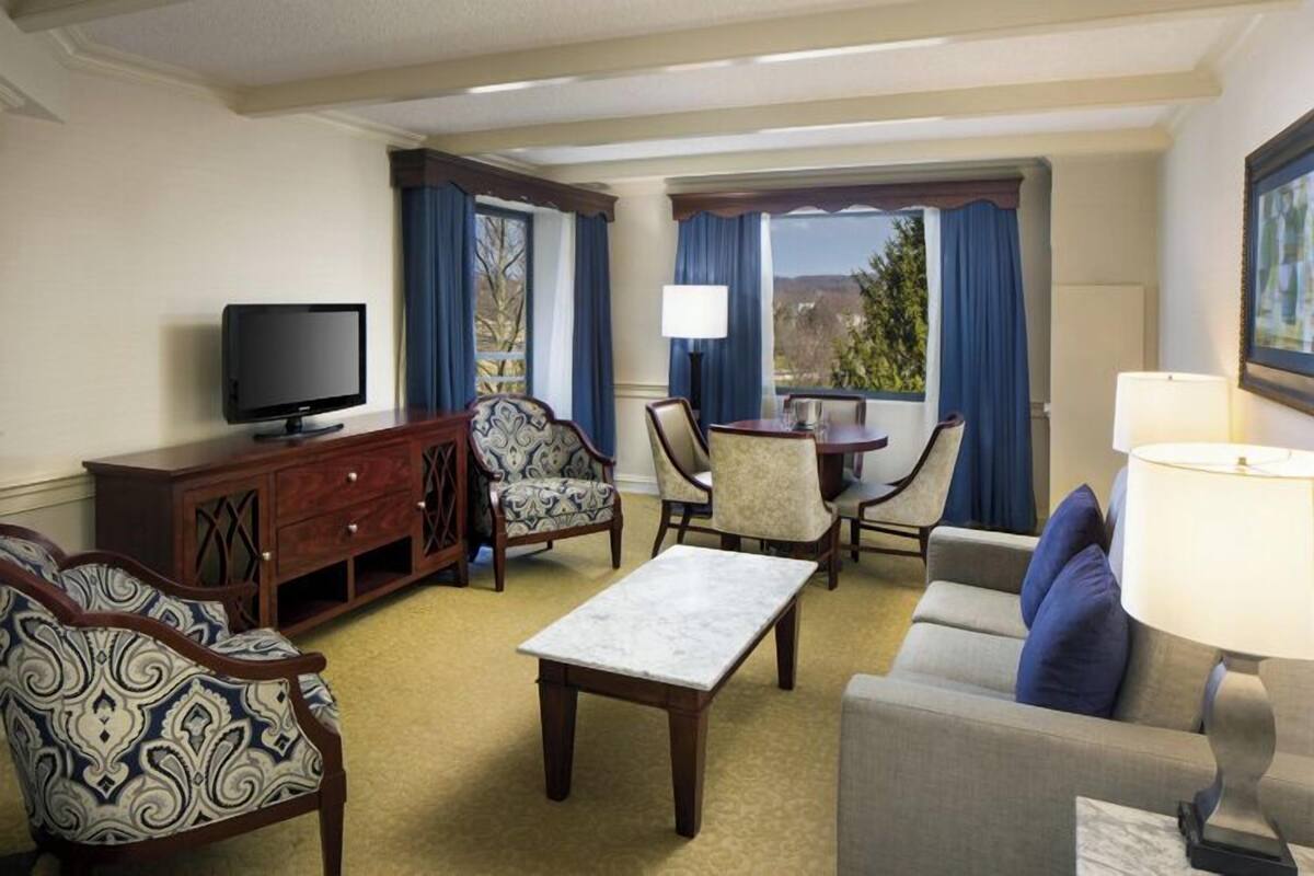 A hotel room with a television, a sofa, a dining table with chairs, and a window with curtains.