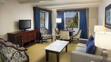 A hotel room with a television, a sofa, a dining table with chairs, and a window with curtains.