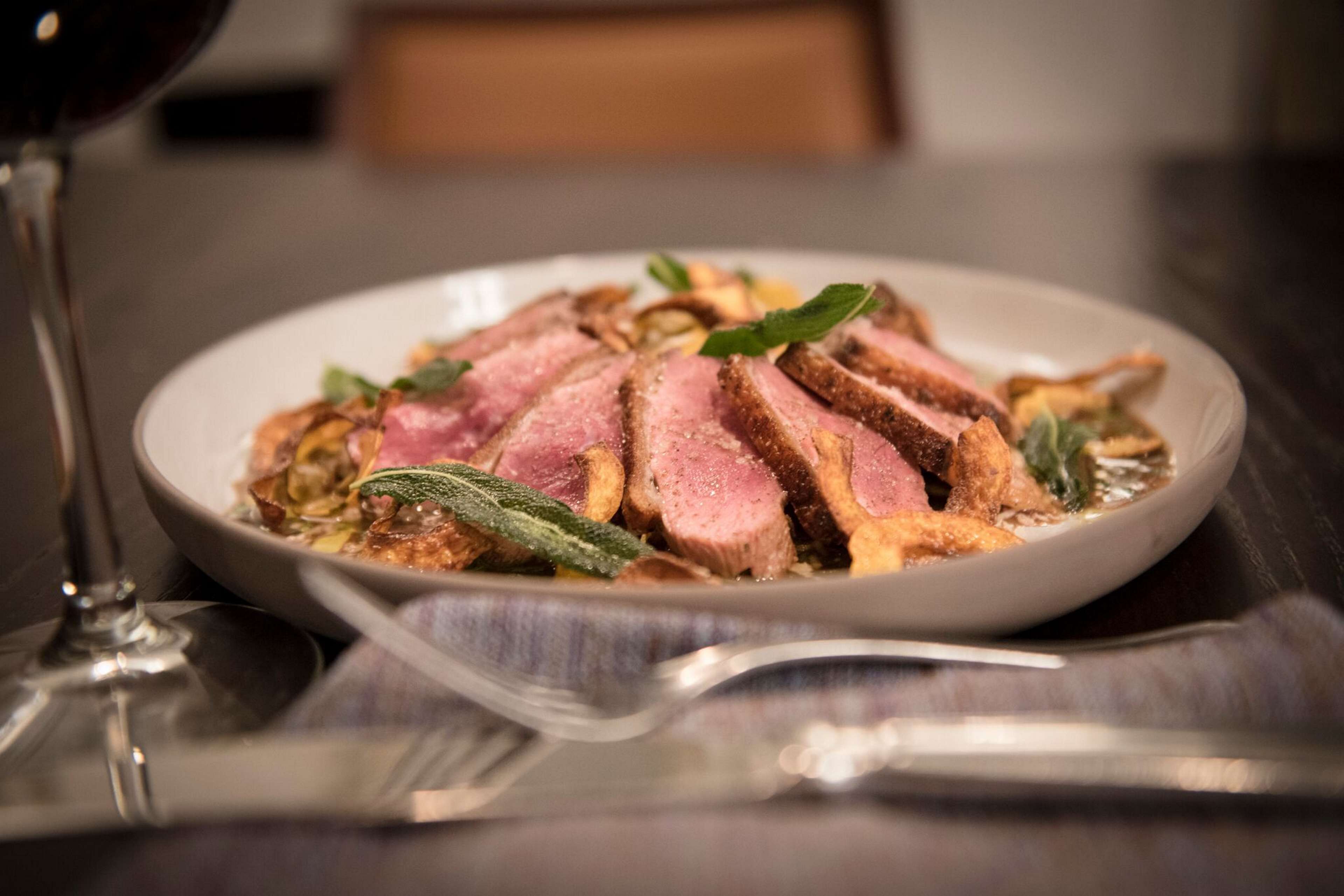 A plate of sliced meat with vegetables and herbs, accompanied by a wine glass and cutlery on a table.