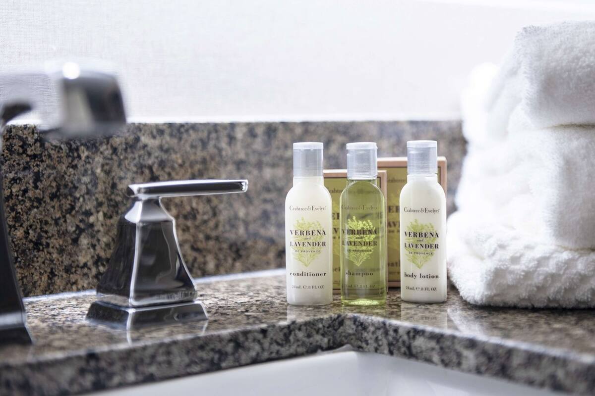 A bathroom counter with a faucet, three bottles of Verbenia products, and a stack of white towels.