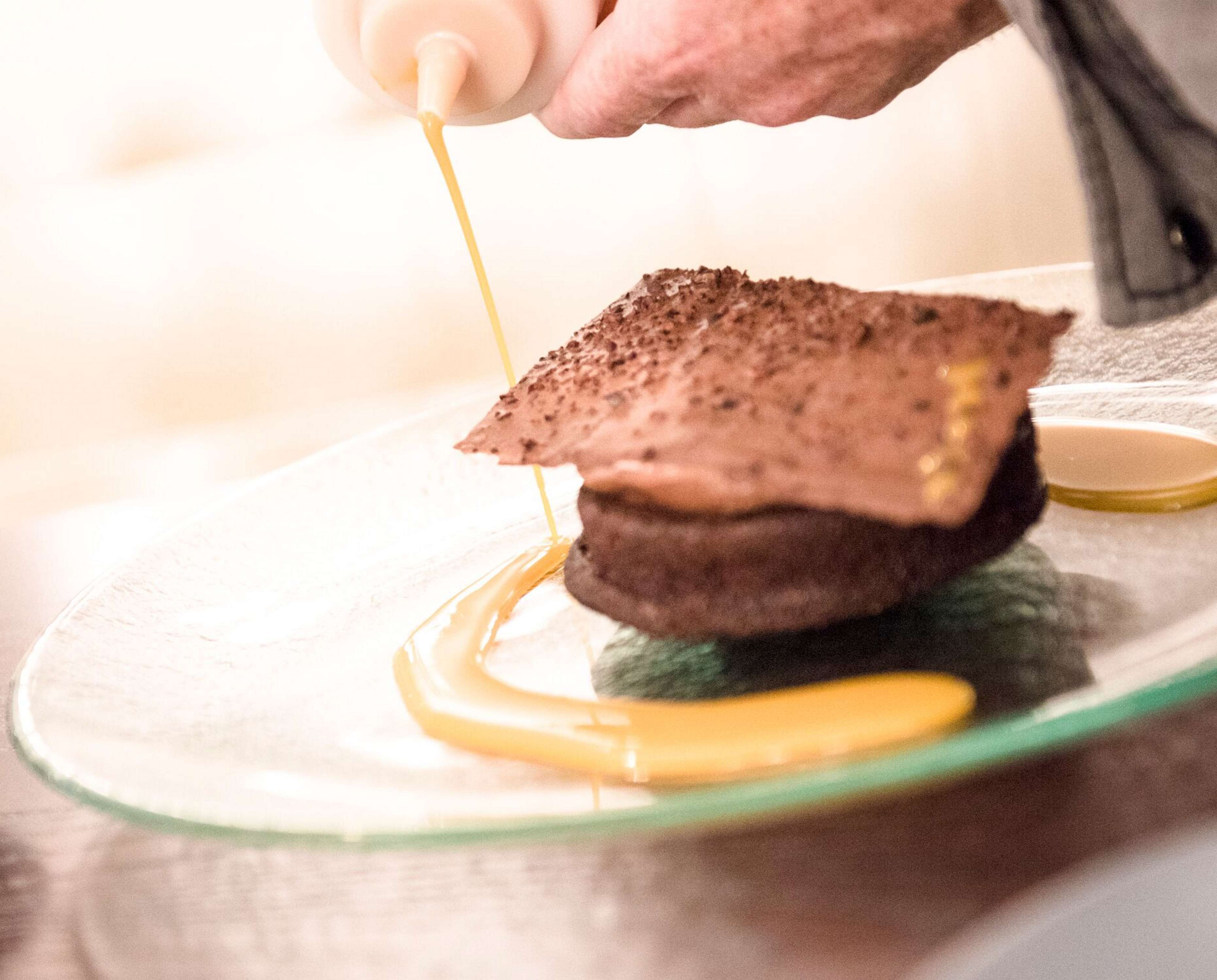 A person pouring a yellow liquid onto a chocolate dessert on a glass plate.