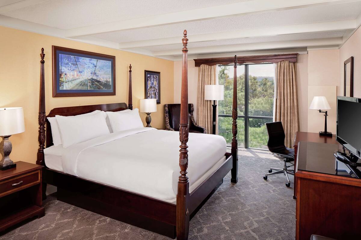A four-poster bed with a cityscape painting, a desk with a TV, and a view of greenery through the window.