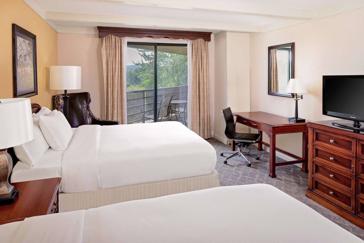 A hotel room with two beds, a desk, a television, and a balcony with a chair.