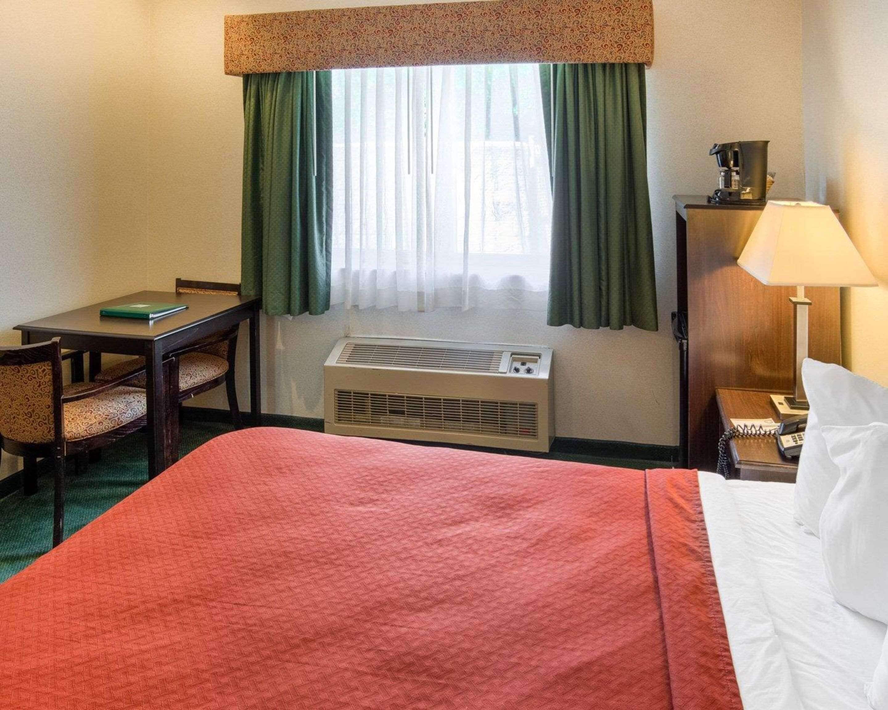 Quarto de hotel com uma cama, uma escrivaninha com cadeira, cafeteira, luminária e ar condicionado.