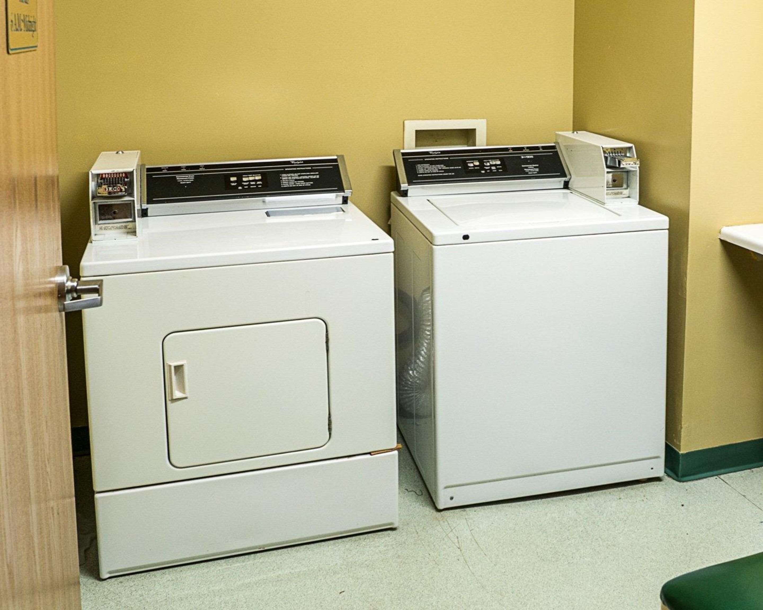 Two stacked front-loading washers in a laundry room with a wooden door and a green bench.