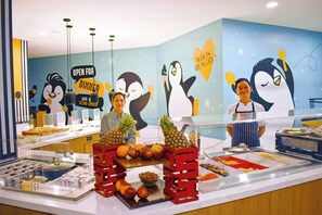 Zona de buffet con una pared temática de pingüinos, diferentes opciones de comida y personal.