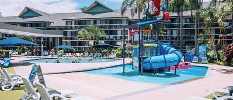 A hotel pool area with a water slide, lounge chairs, and a building with balconies.
