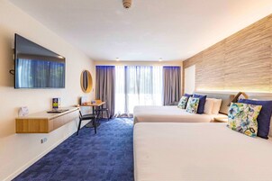 Habitación de hotel con dos camas, un escritorio, una silla, un televisor de pantalla plana y un ventanal grande con cortinas.