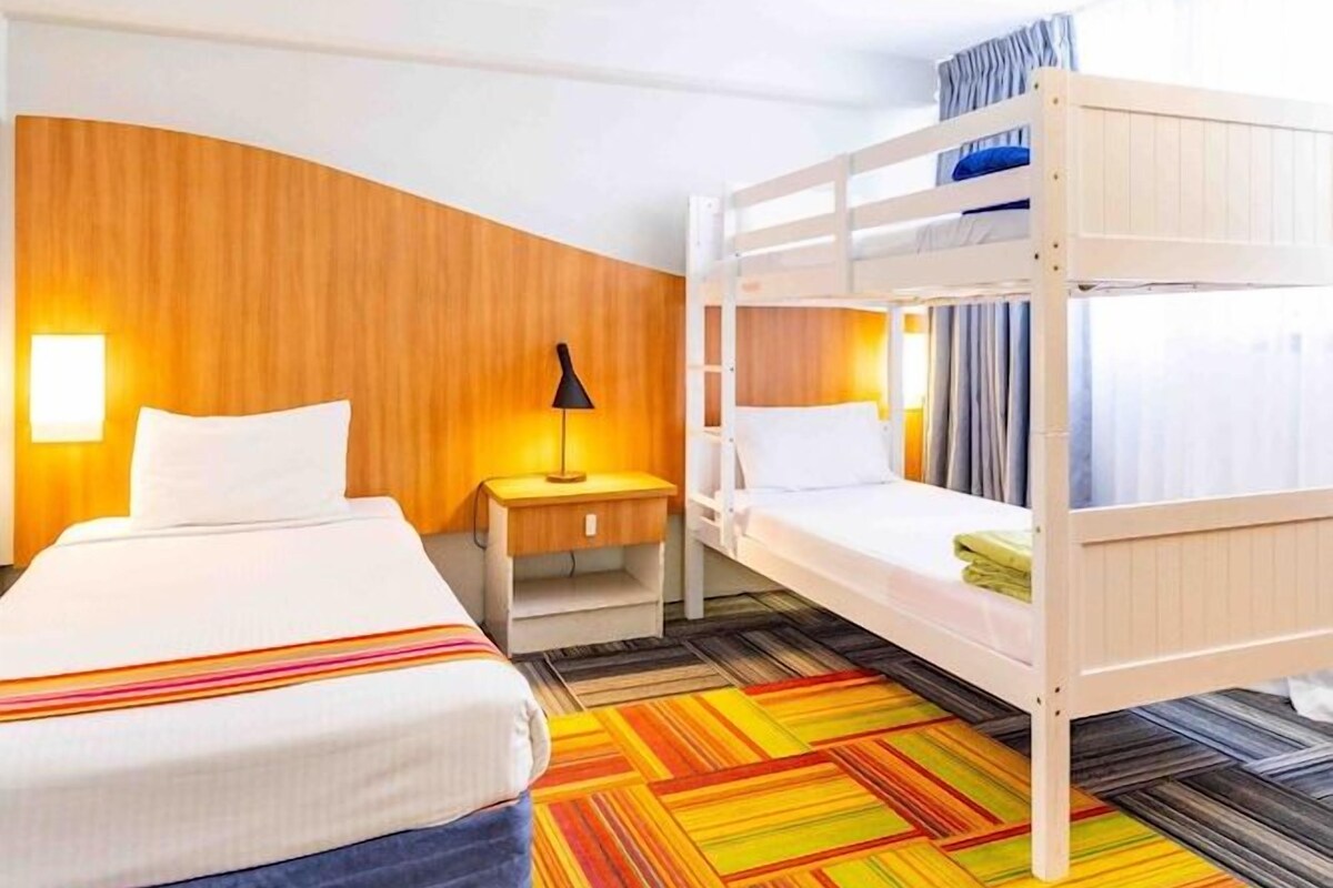 Una habitación de hotel con una cama litera, una cama individual, una mesita de noche y una alfombra a rayas de colores.