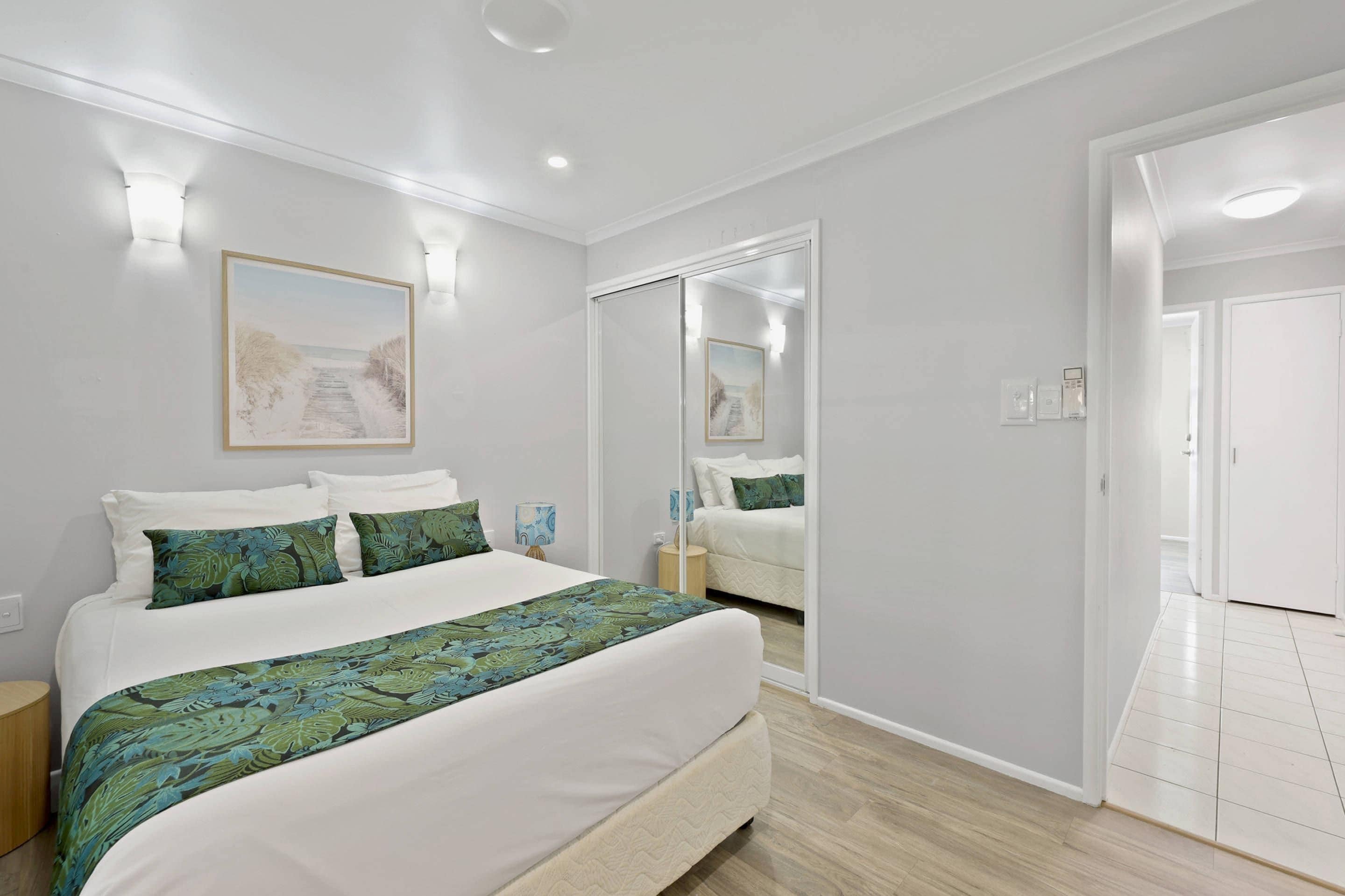 A modern bedroom with a large bed, a bedside table, a nightstand, a mirror, and a door leading to another room.