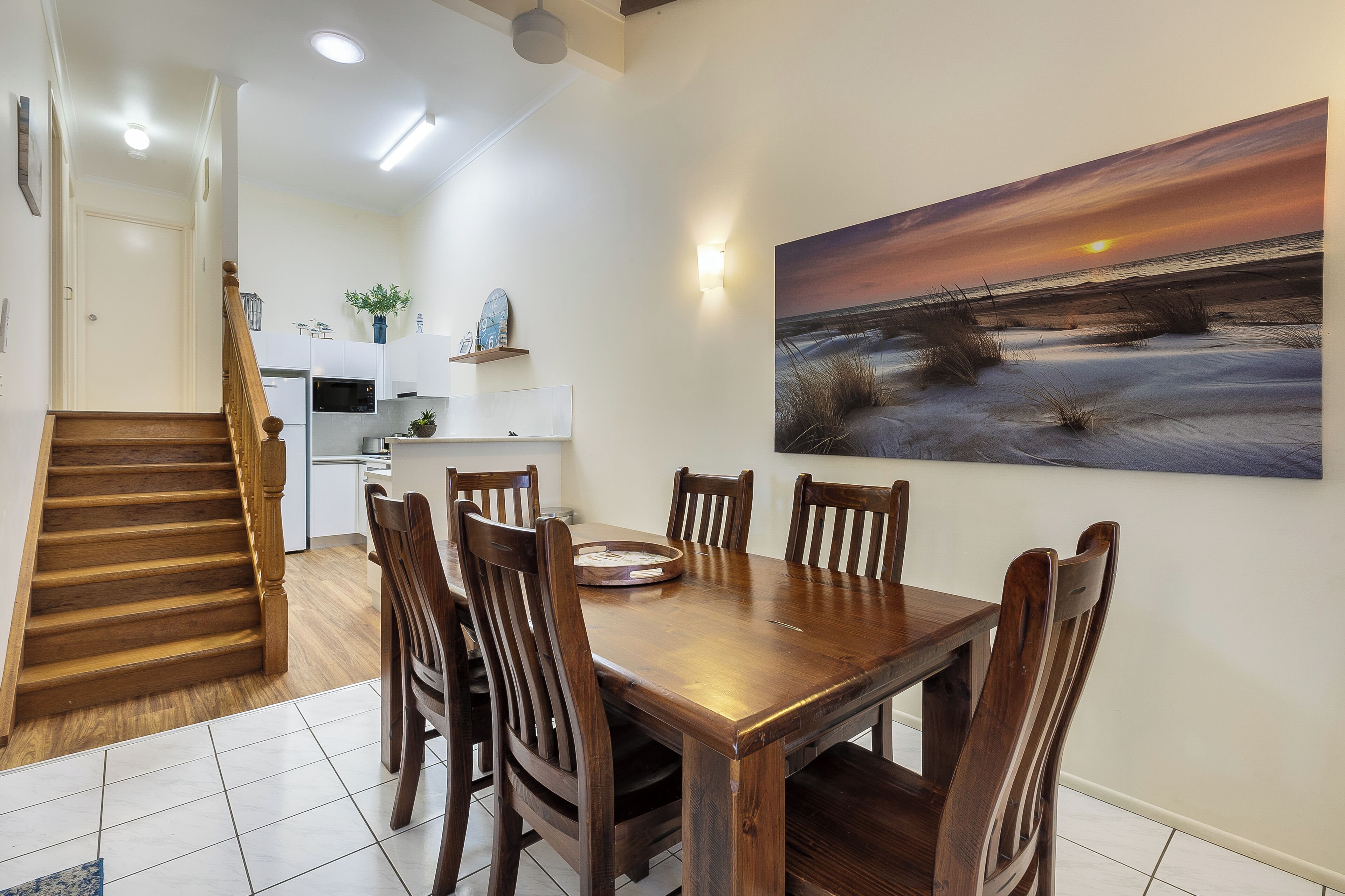 Zona pranzo con tavolo e sedie in legno, cucina sullo sfondo e un quadro raffigurante un tramonto al muro.