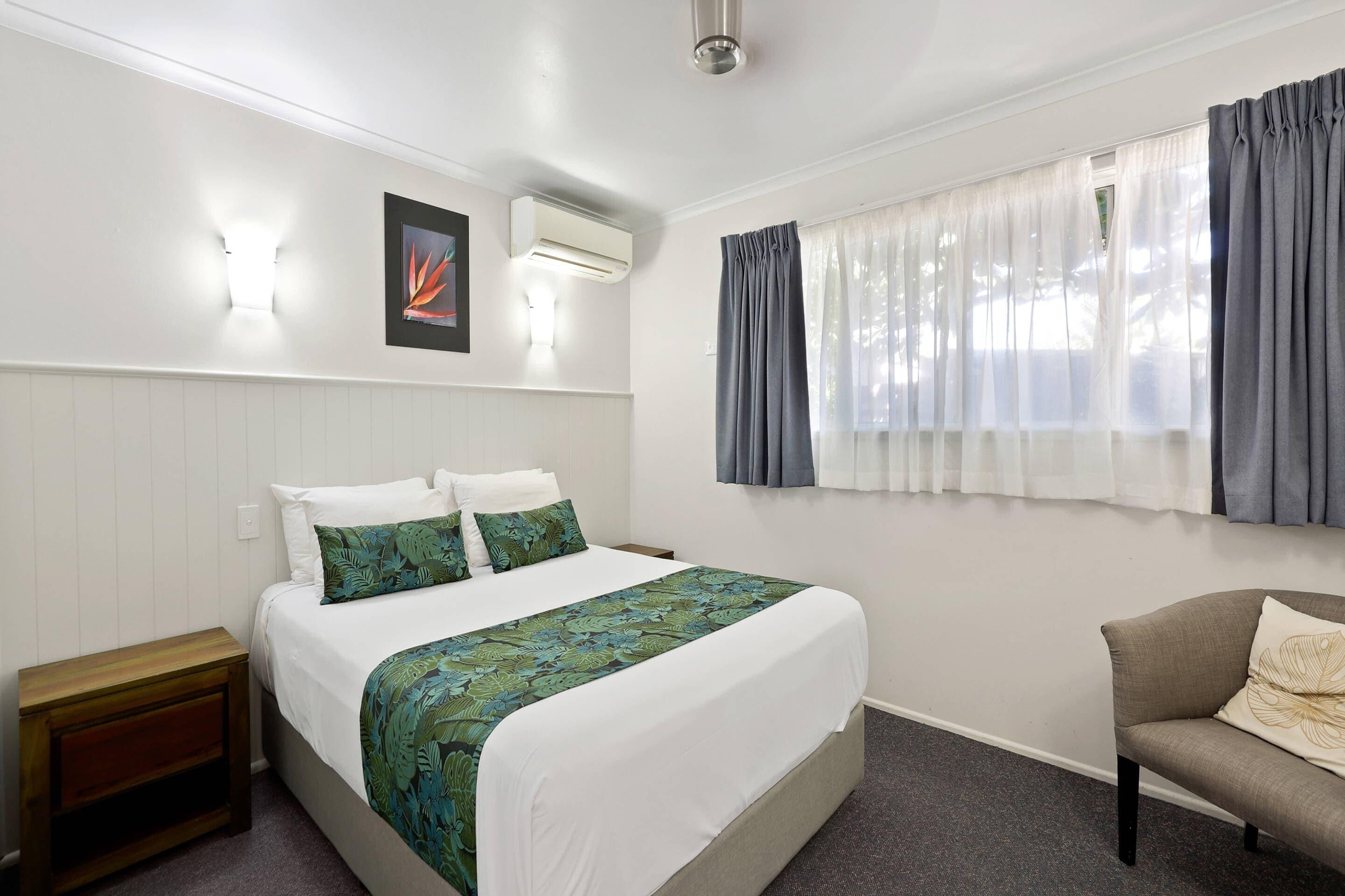 A hotel room with a bed, a nightstand, a chair, a window with curtains, and wall-mounted lighting.