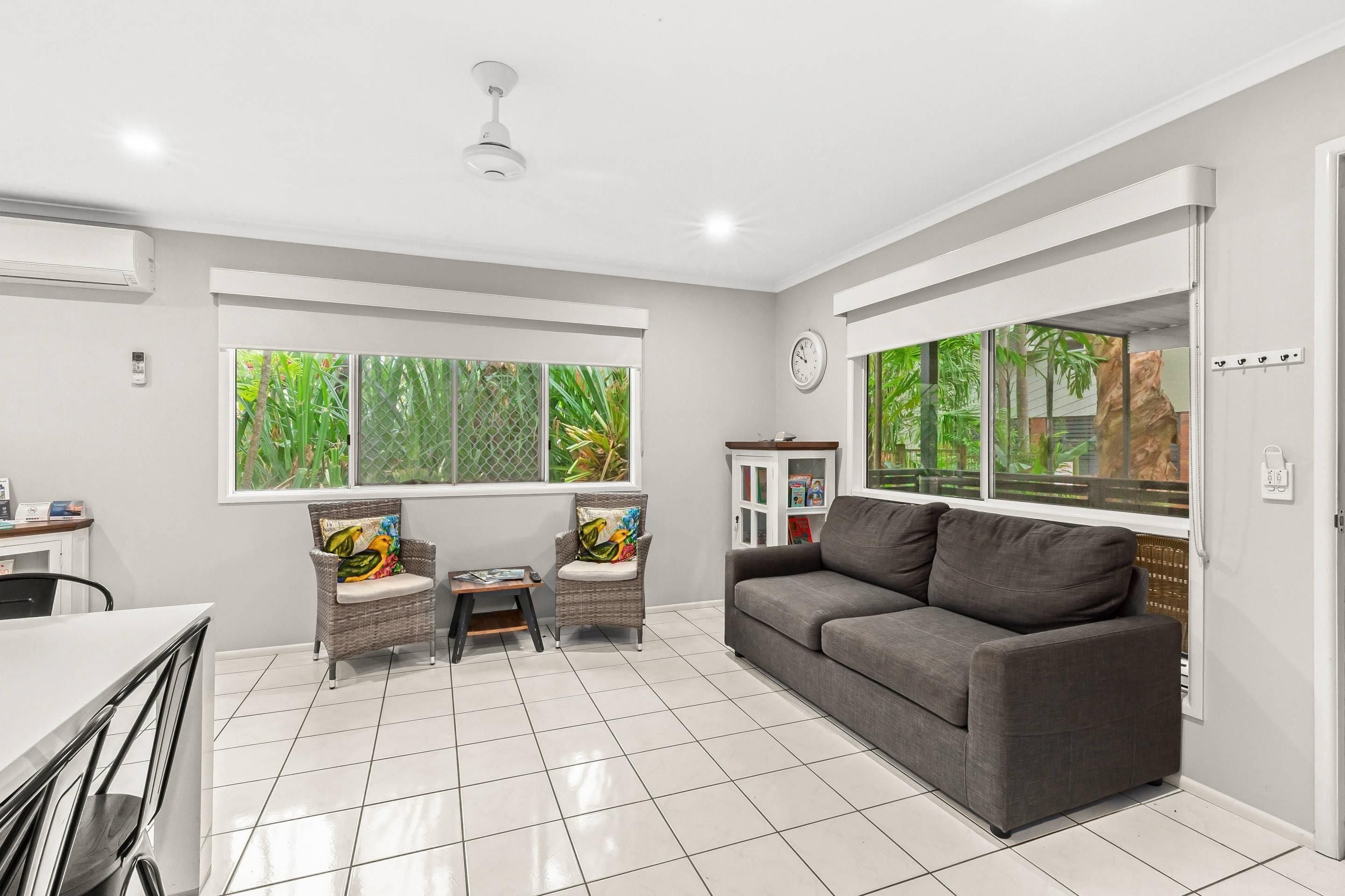 A modern living room with a grey sofa, two wicker chairs, a small table, and large windows with a view of greenery.