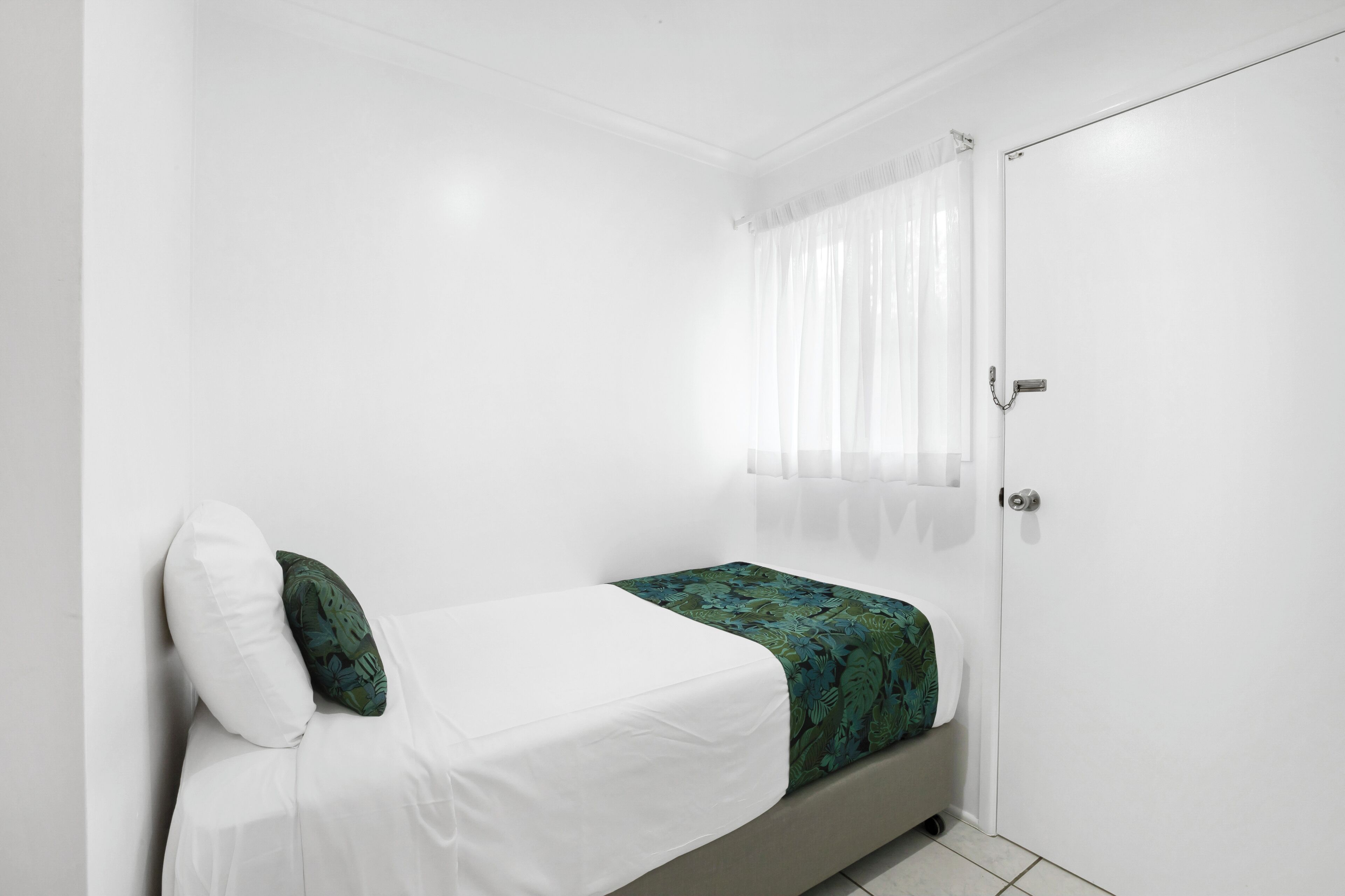 A small, white room with a single bed, a door, and a window with sheer curtains.