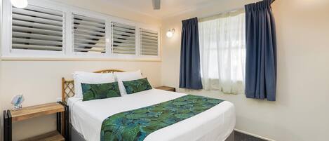 Un dormitorio con cama, mesita de noche, ventilador de techo y ventana con cortinas.