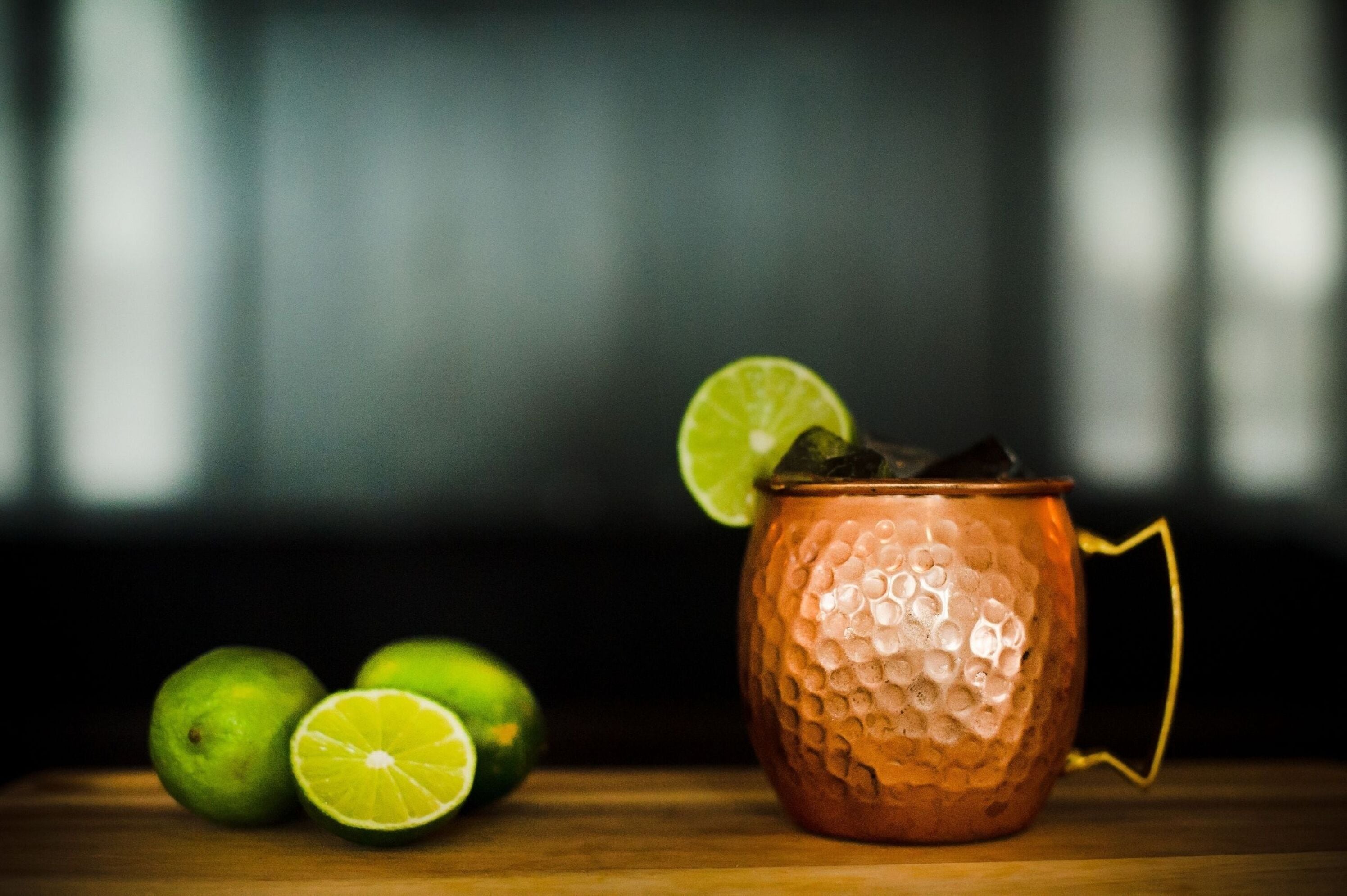 A copper mug with a textured surface and a handle, accompanied by whole and sliced limes on a wooden surface.
