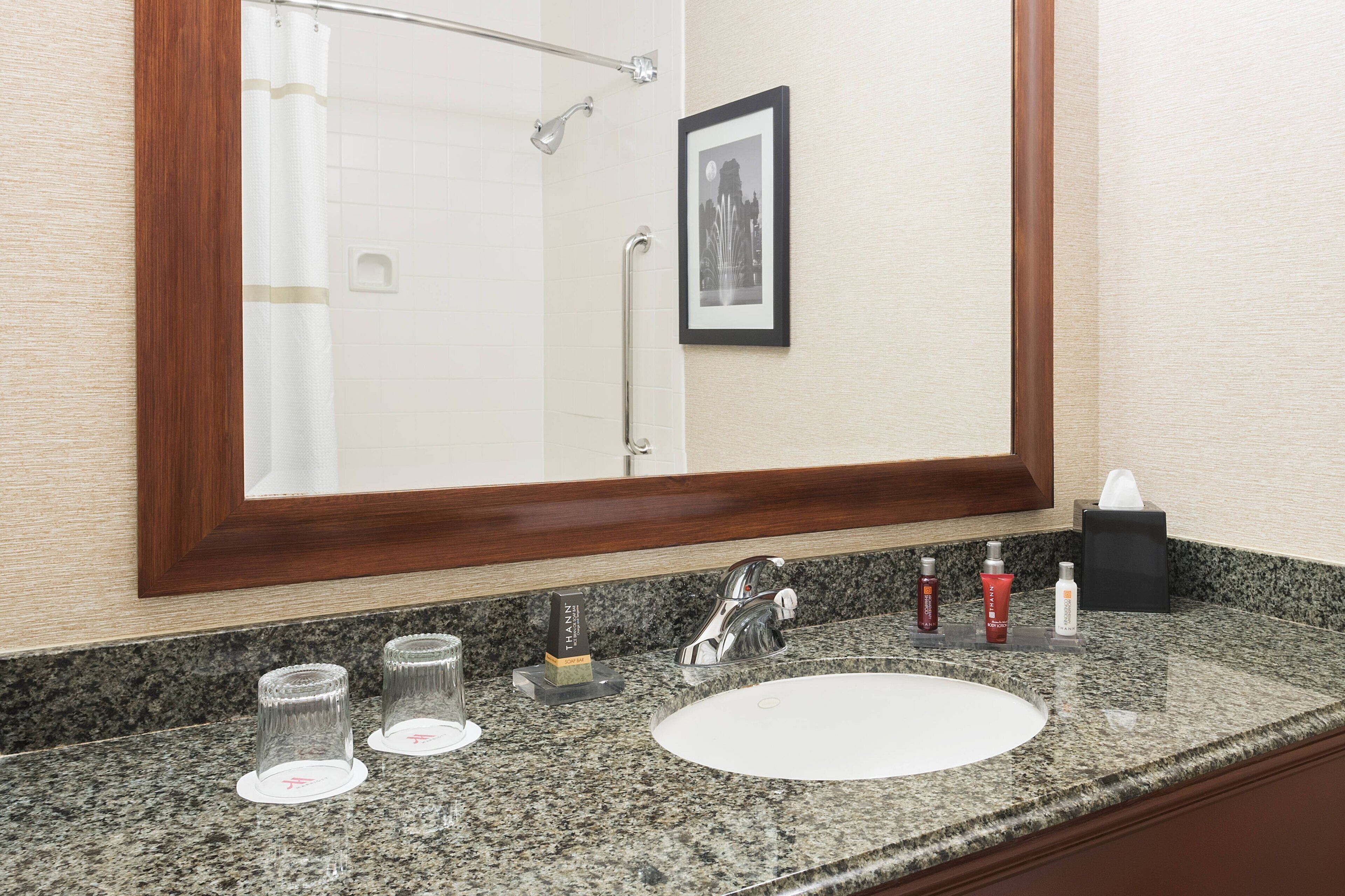 A bathroom with a granite countertop, a white sink, a framed mirror, a shower with a glass door, and a framed picture on the wall.