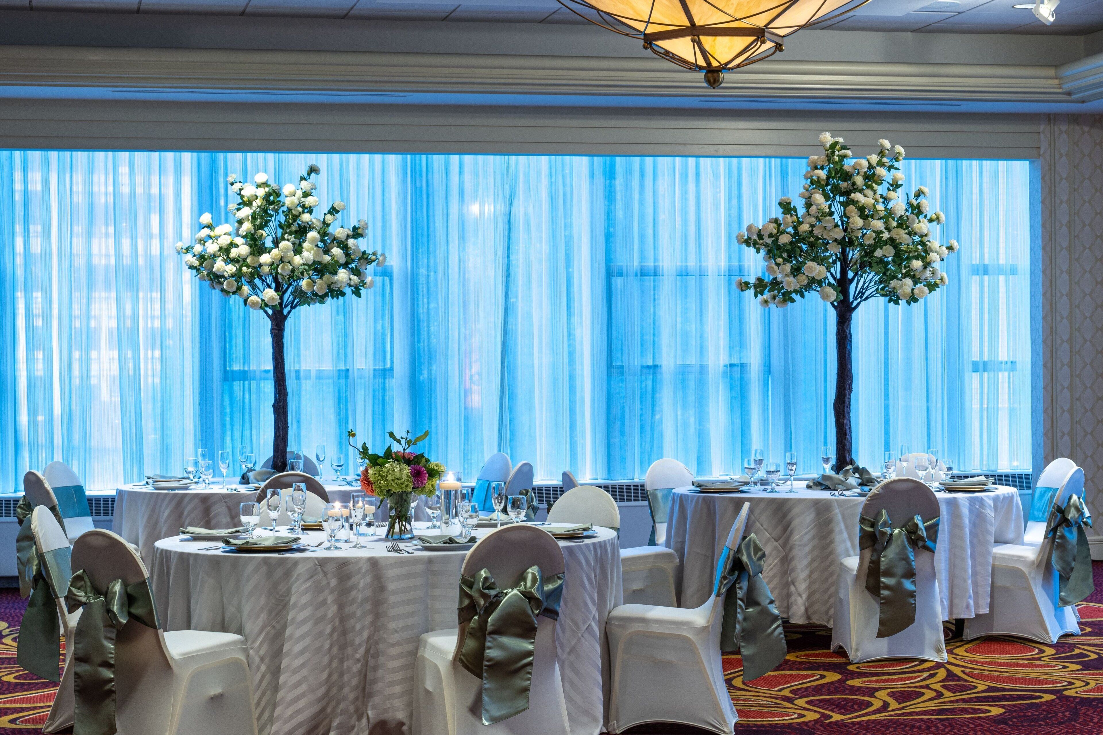 A banquet hall with round tables set for a formal event, featuring white tablecloths, chairs with bows, and floral centerpieces.
