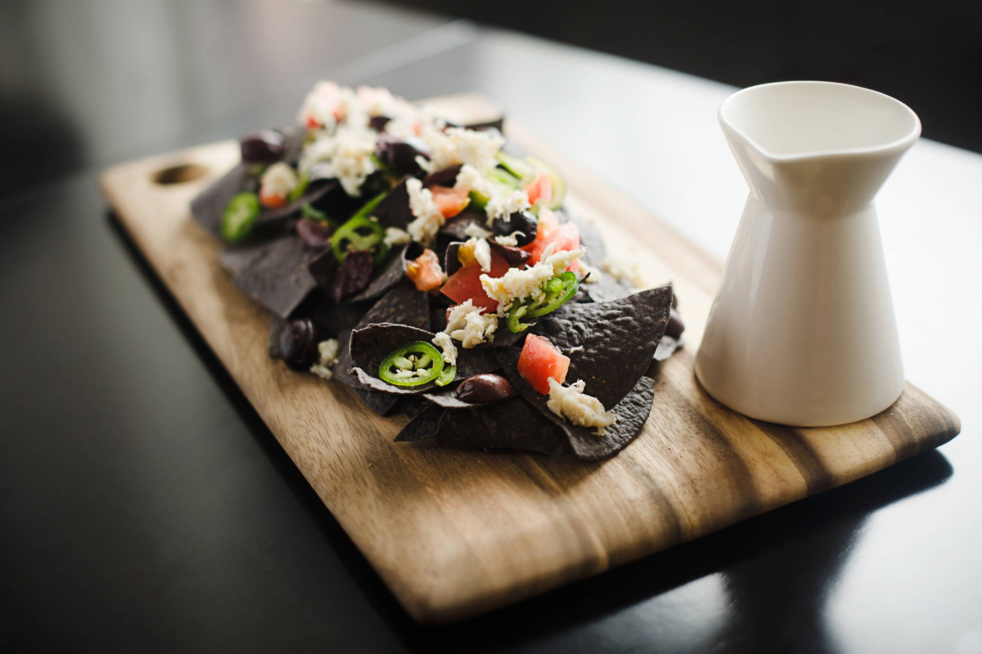 A wooden serving board with a dish of black rice, topped with diced tomatoes, green peppers, and cheese, accompanied by a white pitcher.