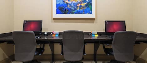 A conference room with two desks, each with a laptop, a phone, and a keyboard. There are three office chairs. A framed abstract painting is on the wall above the desks.