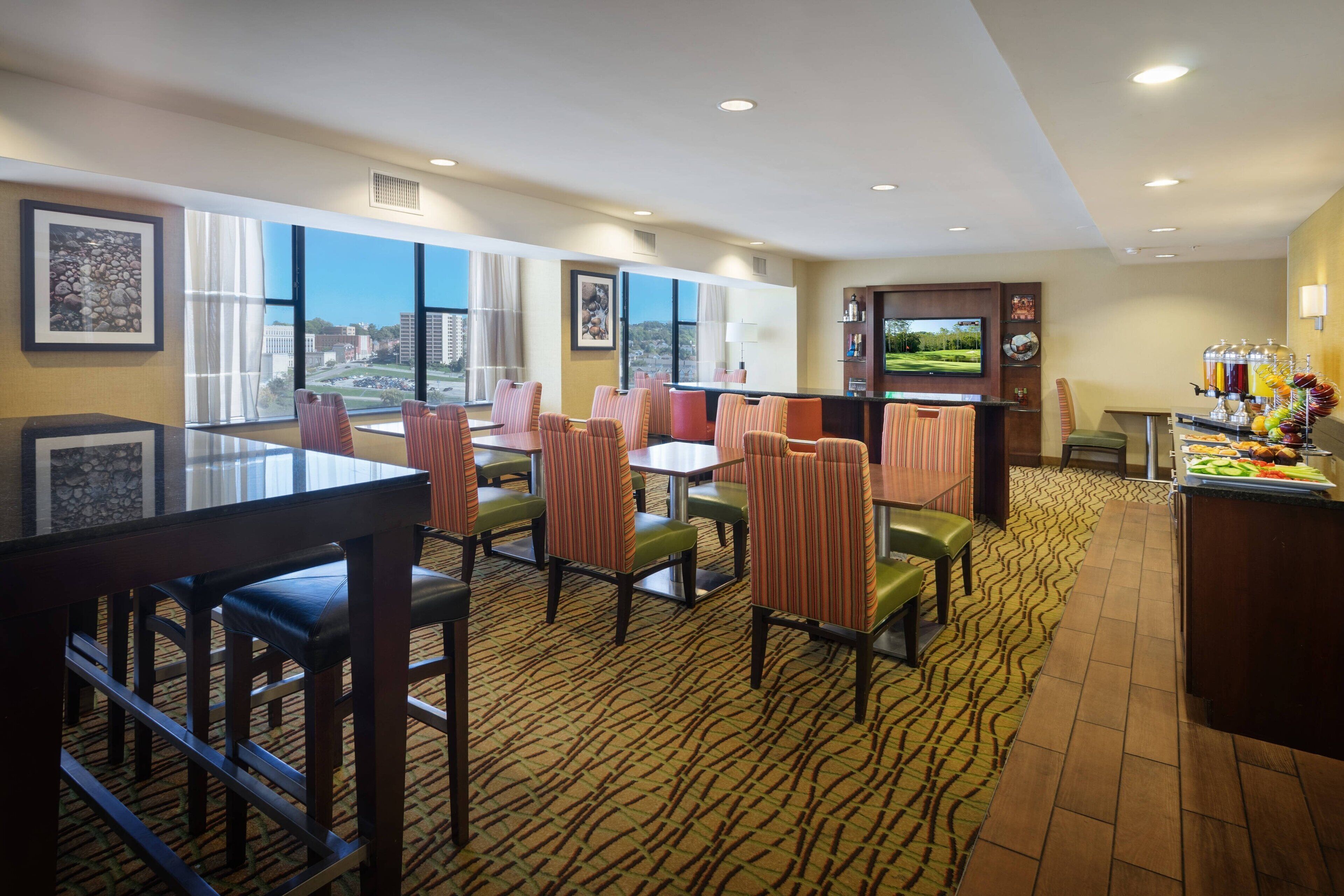 A hotel dining area with multiple tables and chairs, a bar, and a view of the cityscape.