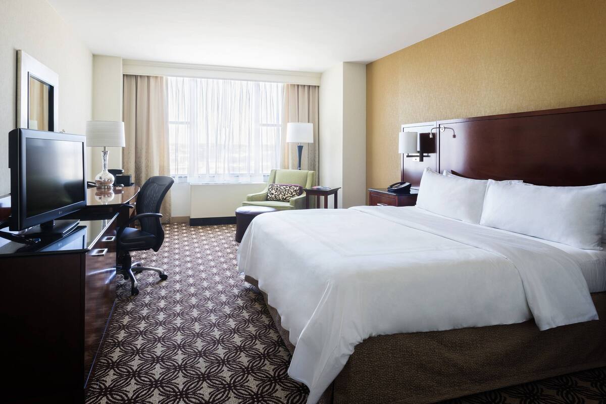 A hotel room with a large bed, a desk with a computer, a chair, a TV, and a window with curtains.