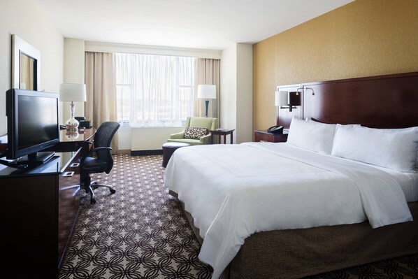 A hotel room with a large bed, a desk with a computer, a chair, a TV, and a window with curtains.