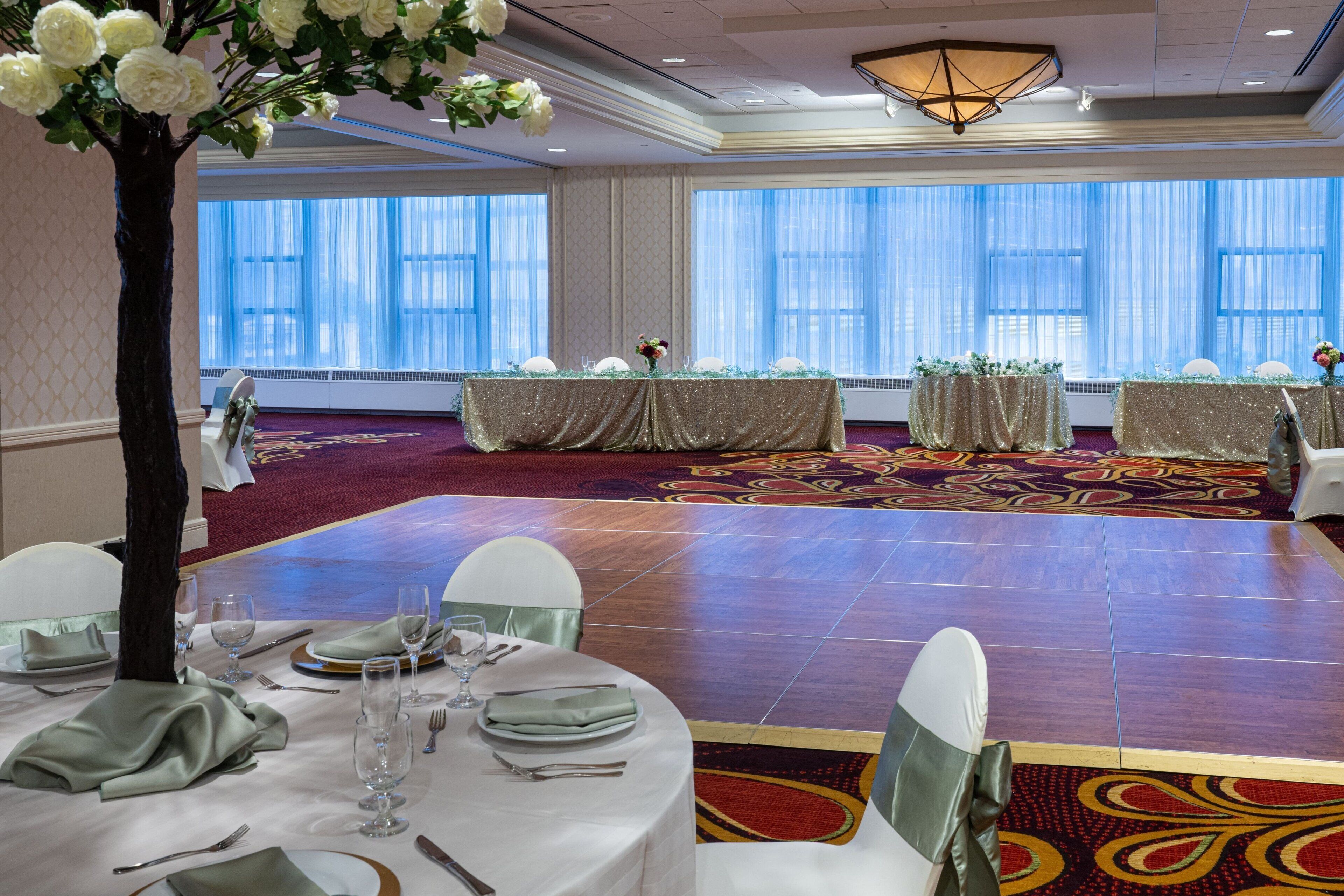 Un salón de banquetes con mesas redondas dispuestas para un evento formal, ventanas amplias con cortinas traslúcidas y un centro de mesa decorativo con un árbol.