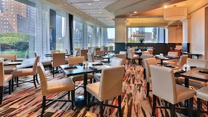 En hotellrestaurang med flera bord och stolar, stora fönster med utsikt över staden och en mönstrad matta.
