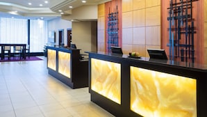 En hotellobby med modern design, med en receptionsdisk med upplysta paneler, en sittgrupp med en bänk och stolar samt en stor vägg med ett geometriskt konstverk.