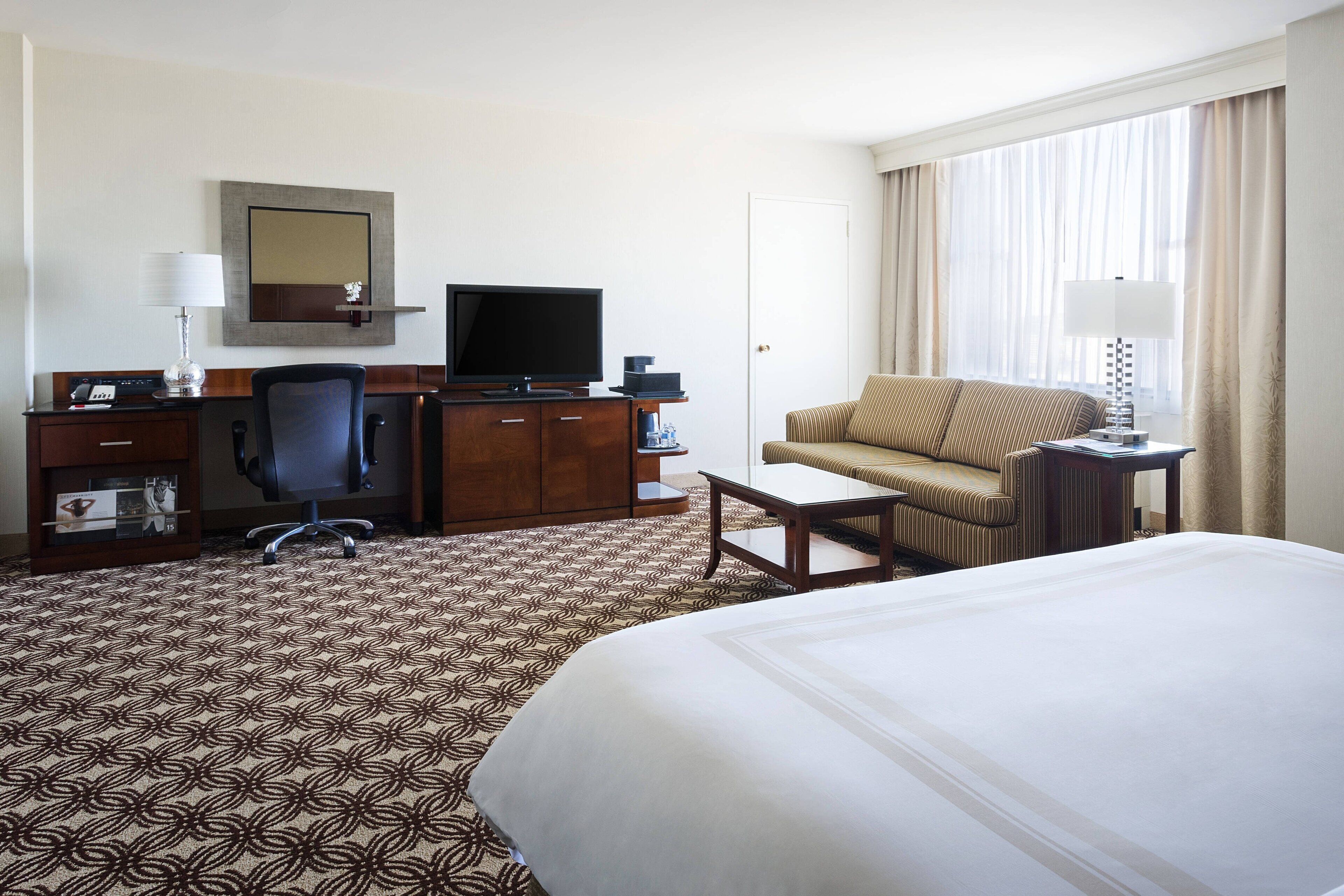 Ein Hotelzimmer mit einem großen Bett, einem Schreibtisch mit Computer, einem Sessel, einem Fernseher und einem Fenster mit Vorhängen.