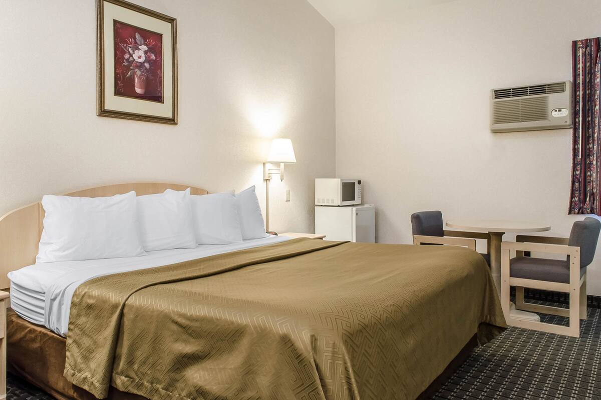A hotel room with a bed, bedside table, lamp, air conditioning unit, and a small desk with a chair.