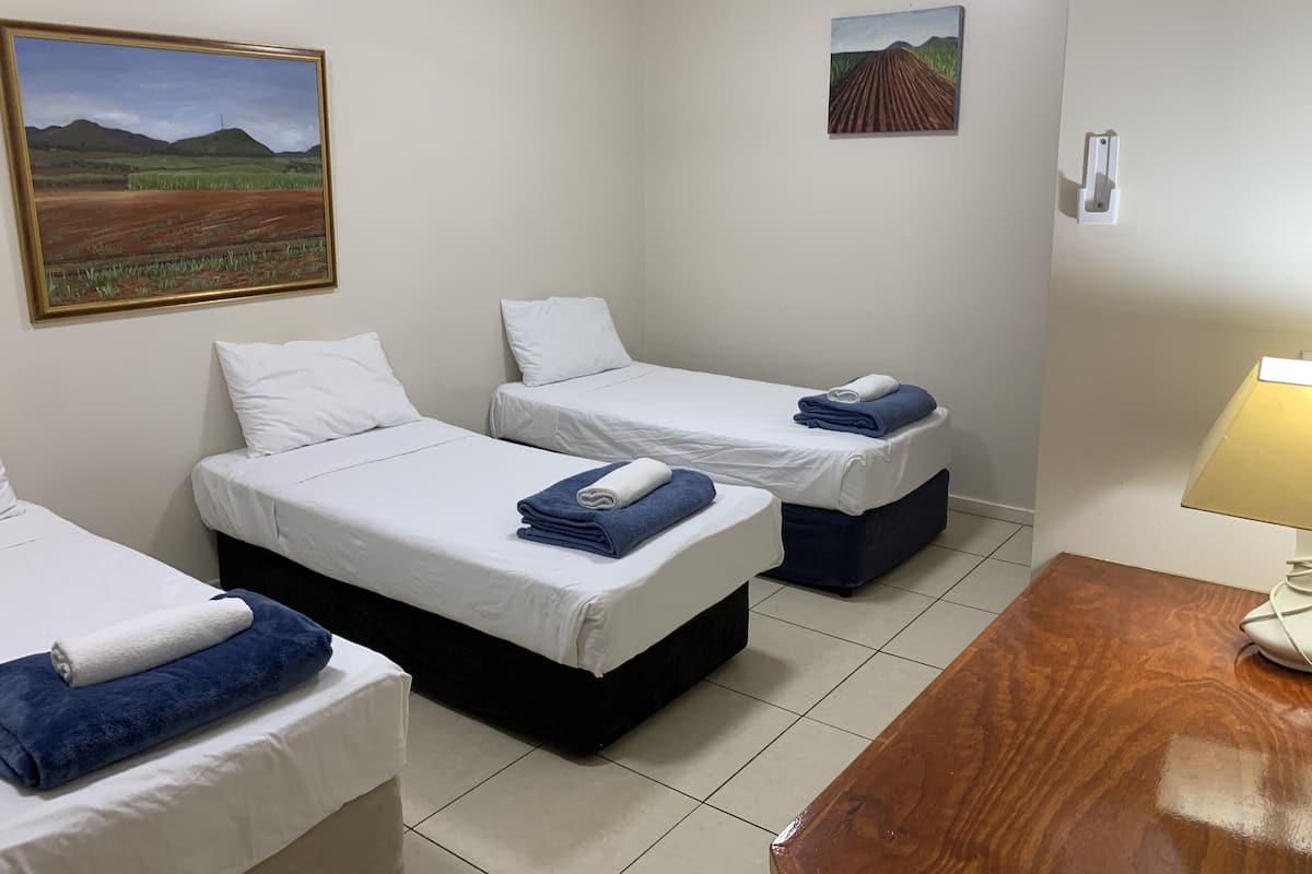 Quarto de hotel com duas camas de solteiro, cada uma com roupa de cama branca e toalhas azuis. Há uma pintura na parede acima das camas, uma mesa pequena com abajur e piso de azulejos.