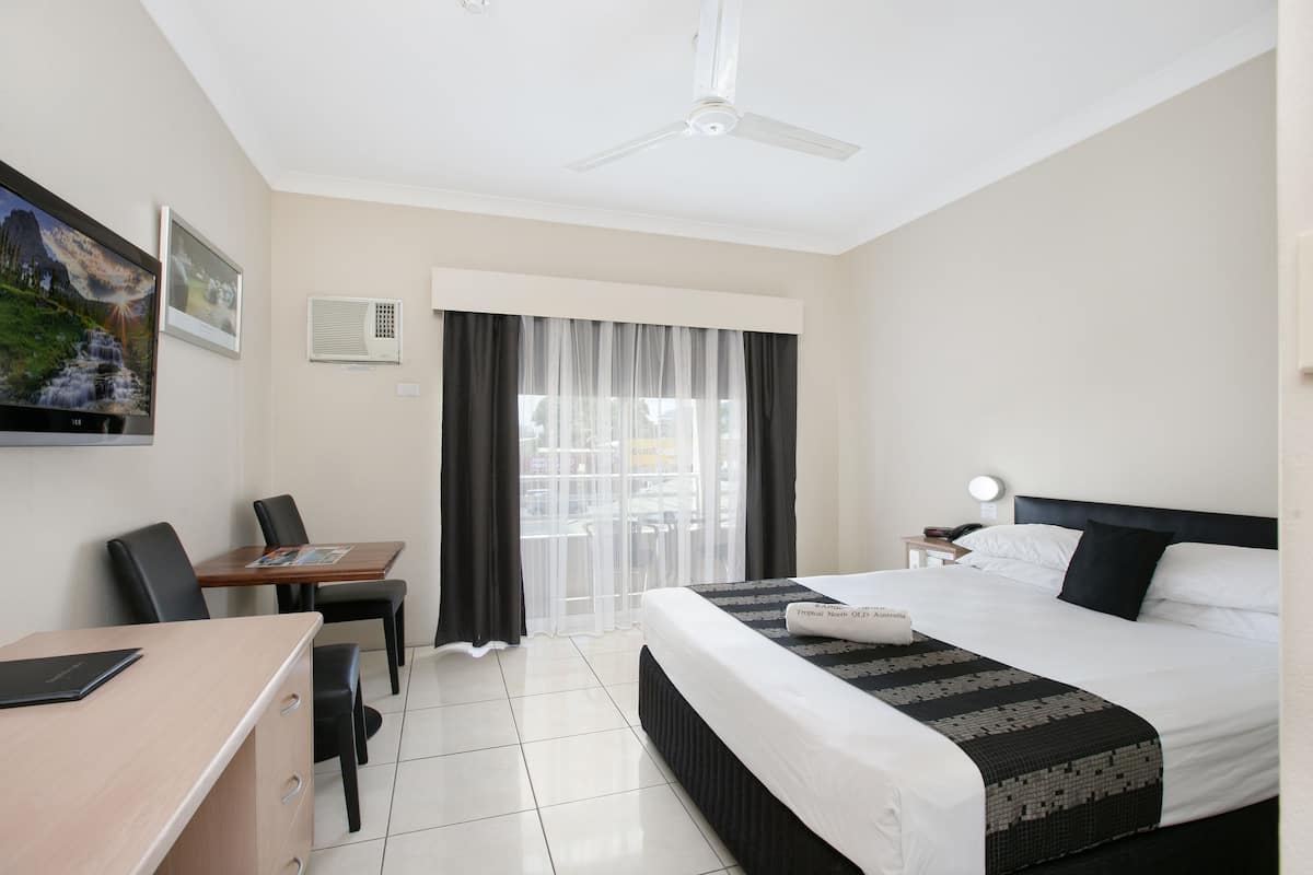 Quarto de hotel com cama, escrivaninha, cadeiras, ventilador de teto e janela com cortinas finas.