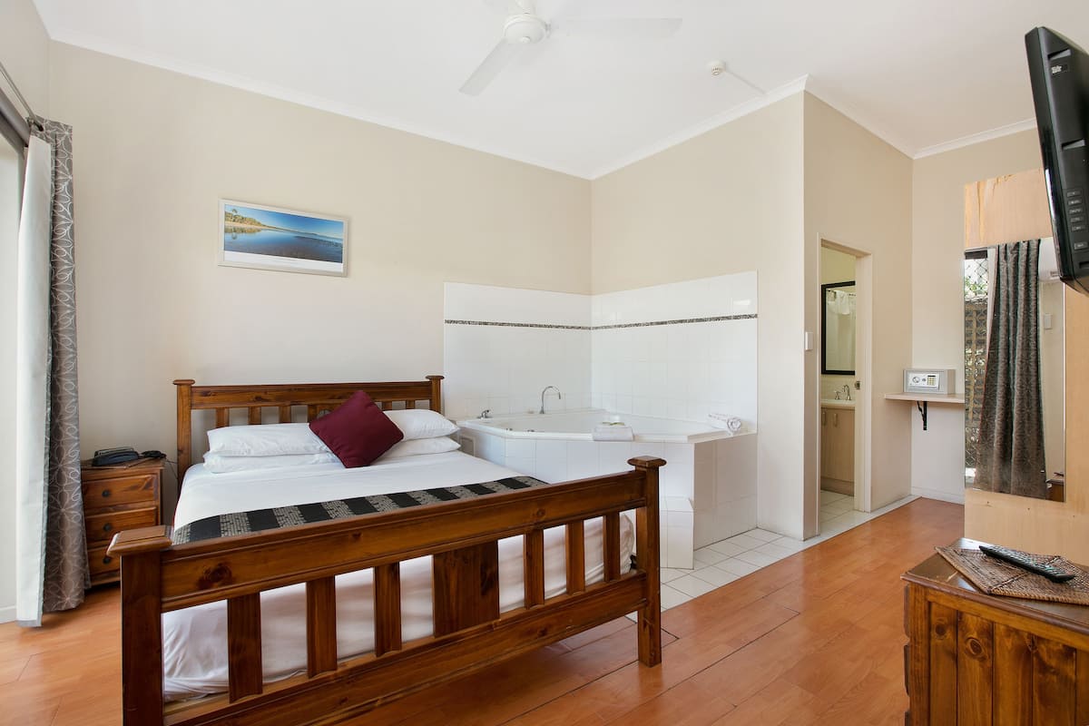 Quarto com cama de madeira, banheira branca e televisão sobre suporte de madeira.