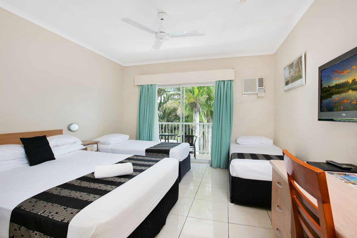 Quarto de hotel com duas camas, uma escrivaninha, uma cadeira, um ventilador de teto, uma janela com cortinas e um quadro na parede.
