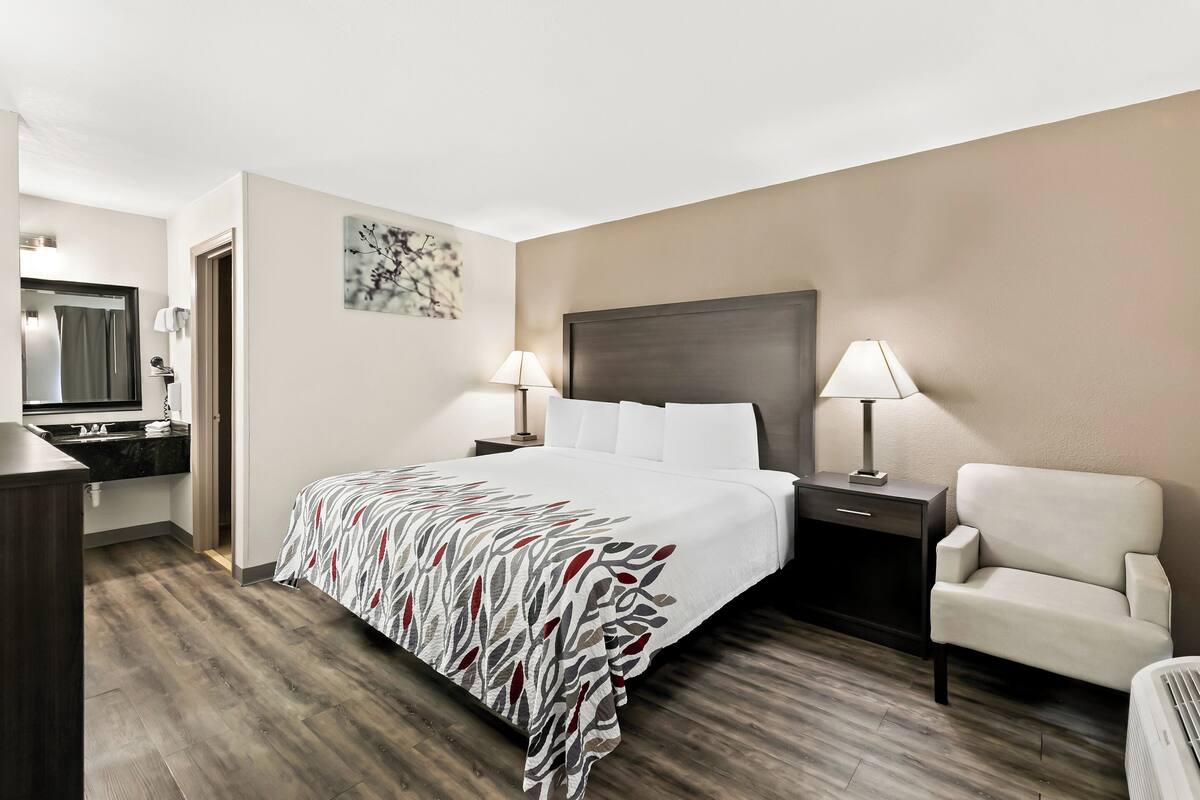 Ein Hotelzimmer mit einem Bett, zwei Nachttischen mit Lampen, einem Sessel, einem Schreibtisch mit Spiegel und einem Wandbild.