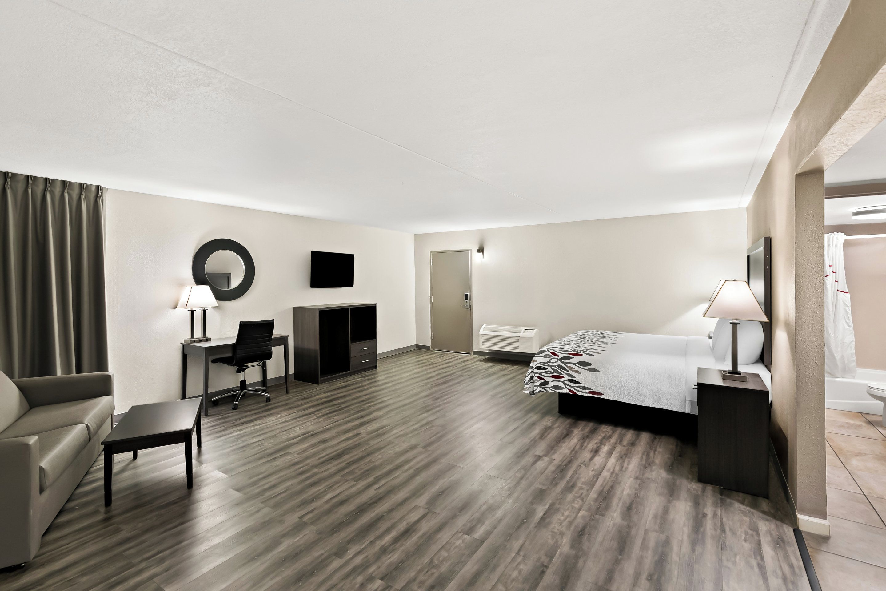 A hotel room with a bed, a desk, a chair, a sofa, a small table, a mirror, and a TV.