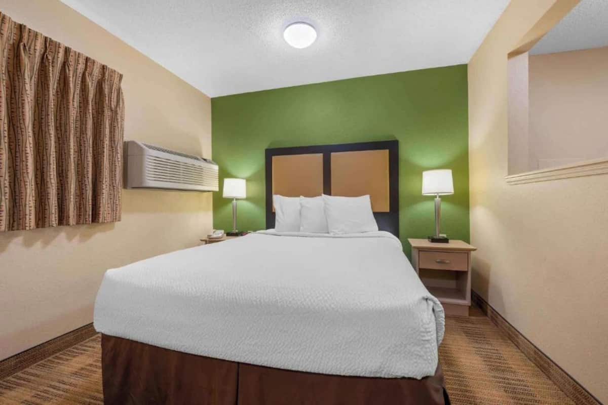 A hotel room with a large bed, two bedside tables with lamps, a wall-mounted air conditioner, and a green accent wall.