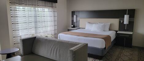 A hotel room with a bed, a sofa, a bedside table, a lamp, and a window with curtains.