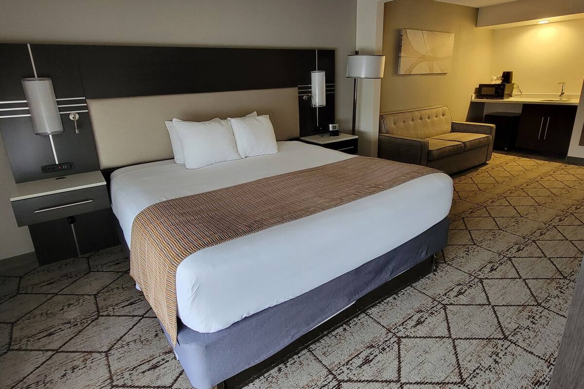 A hotel room with a large bed, a bedside table, a lamp, a chair, and a kitchenette area.