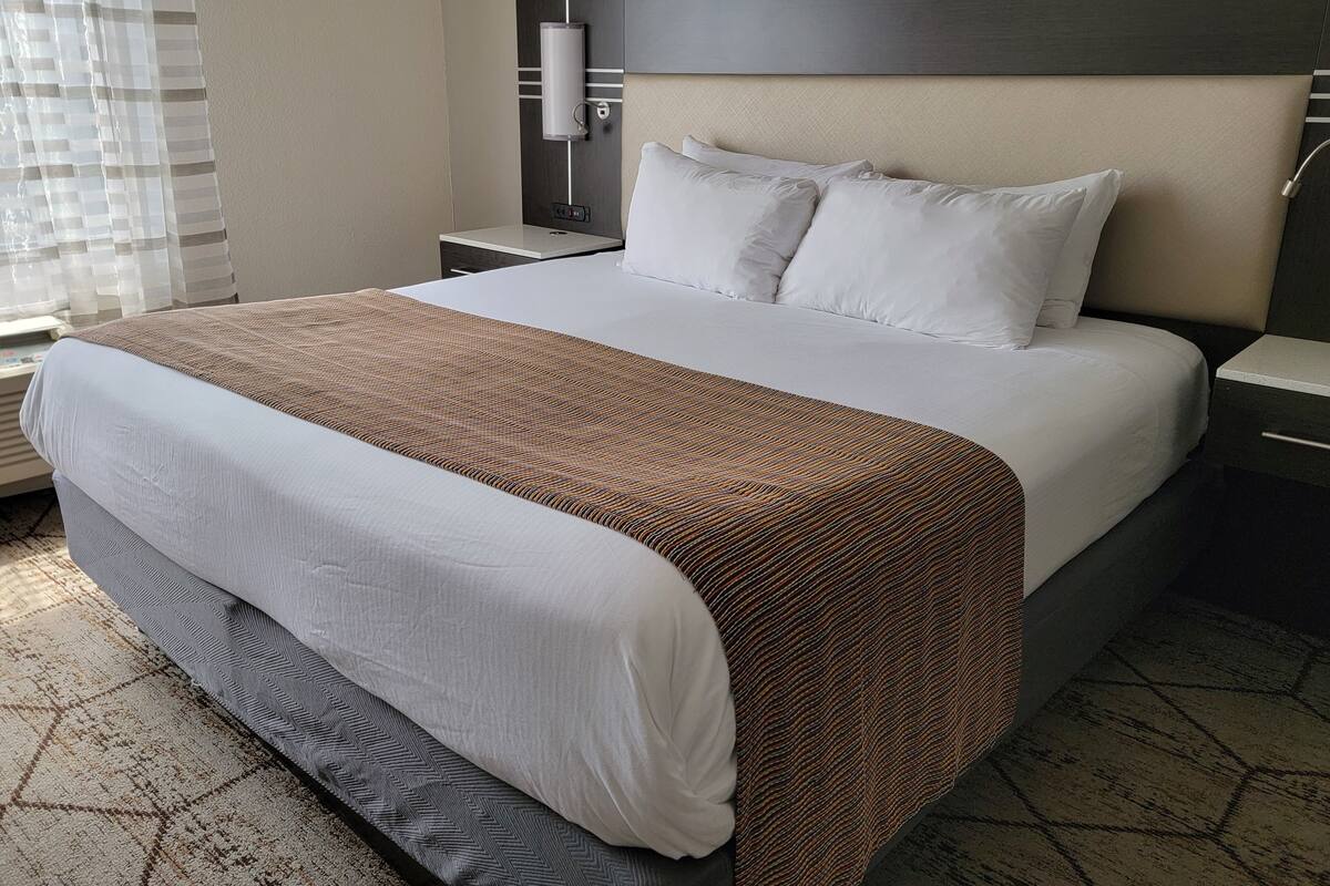 A hotel room with a large bed, a headboard with a light fixture, a bedside table, and a window with blinds.
