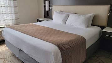 A hotel room with a large bed, a headboard with a light fixture, a bedside table, and a window with blinds.