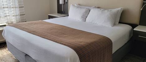 A hotel room with a large bed, a headboard with a light fixture, a bedside table, and a window with blinds.