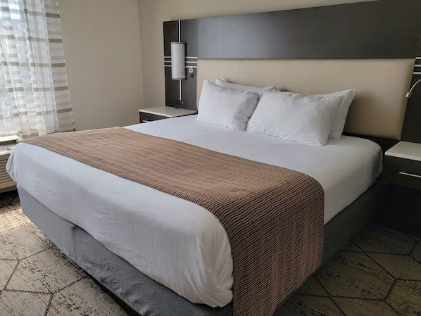 A hotel room with a large bed, a headboard with a light fixture, a bedside table, and a window with blinds.