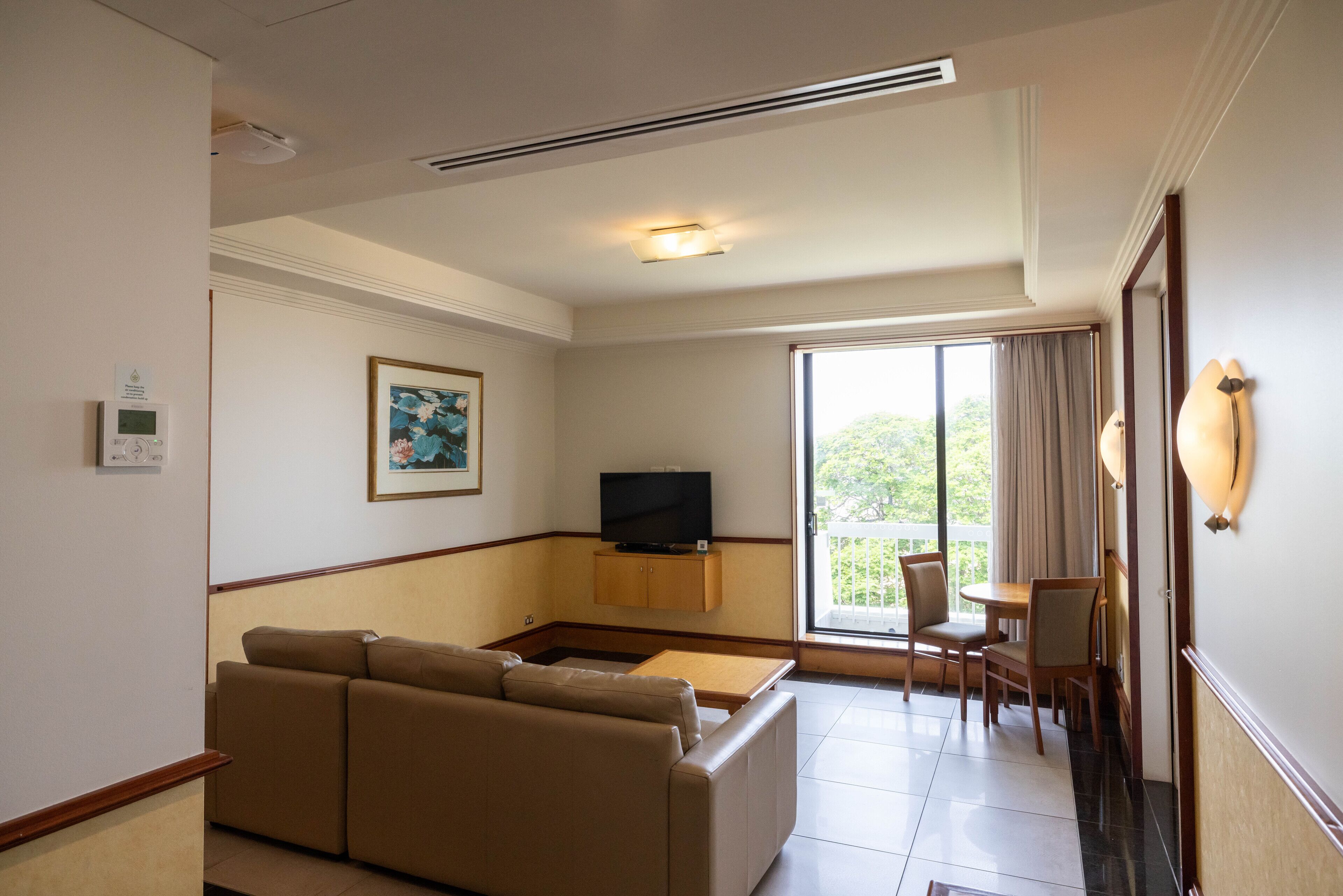 A hotel room with a sofa, a TV, a small table, and a balcony with chairs.