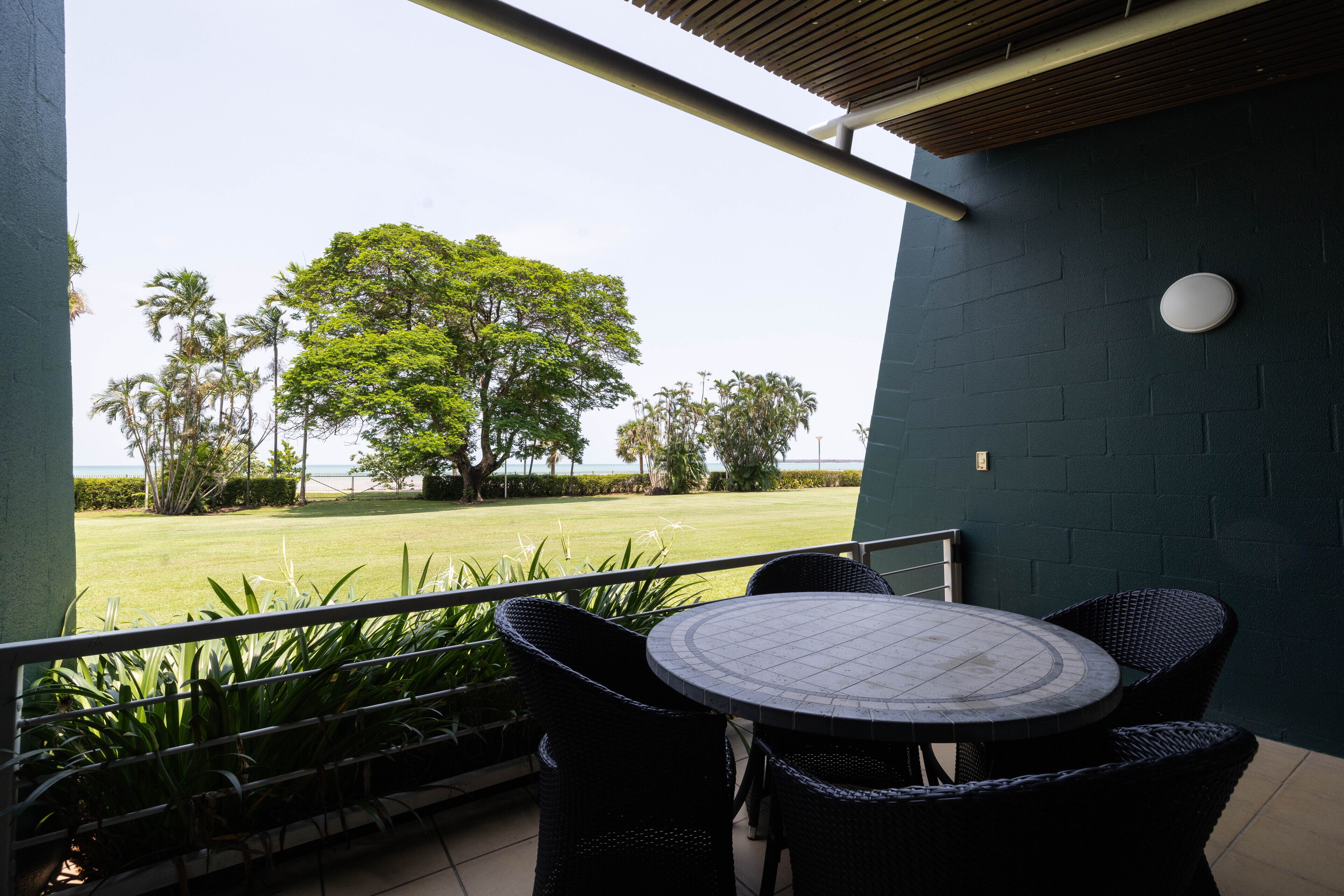 Un patio con una mesa redonda y sillas, vista a un área verde con árboles, y un edificio con pared oscura y un accesorio de iluminación.