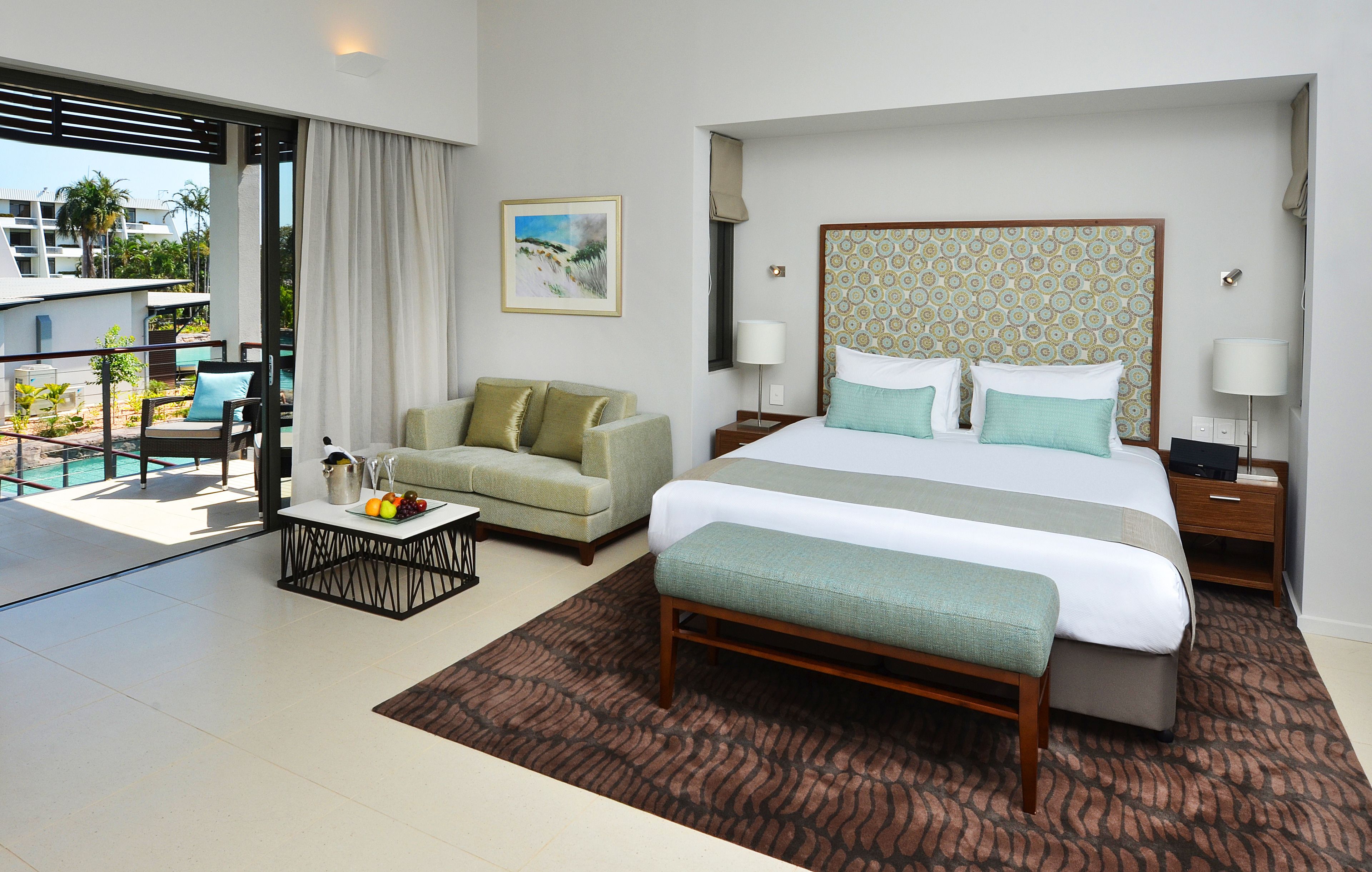 A hotel room with a large bed, a sofa, a coffee table, and a view of the pool and buildings outside.
