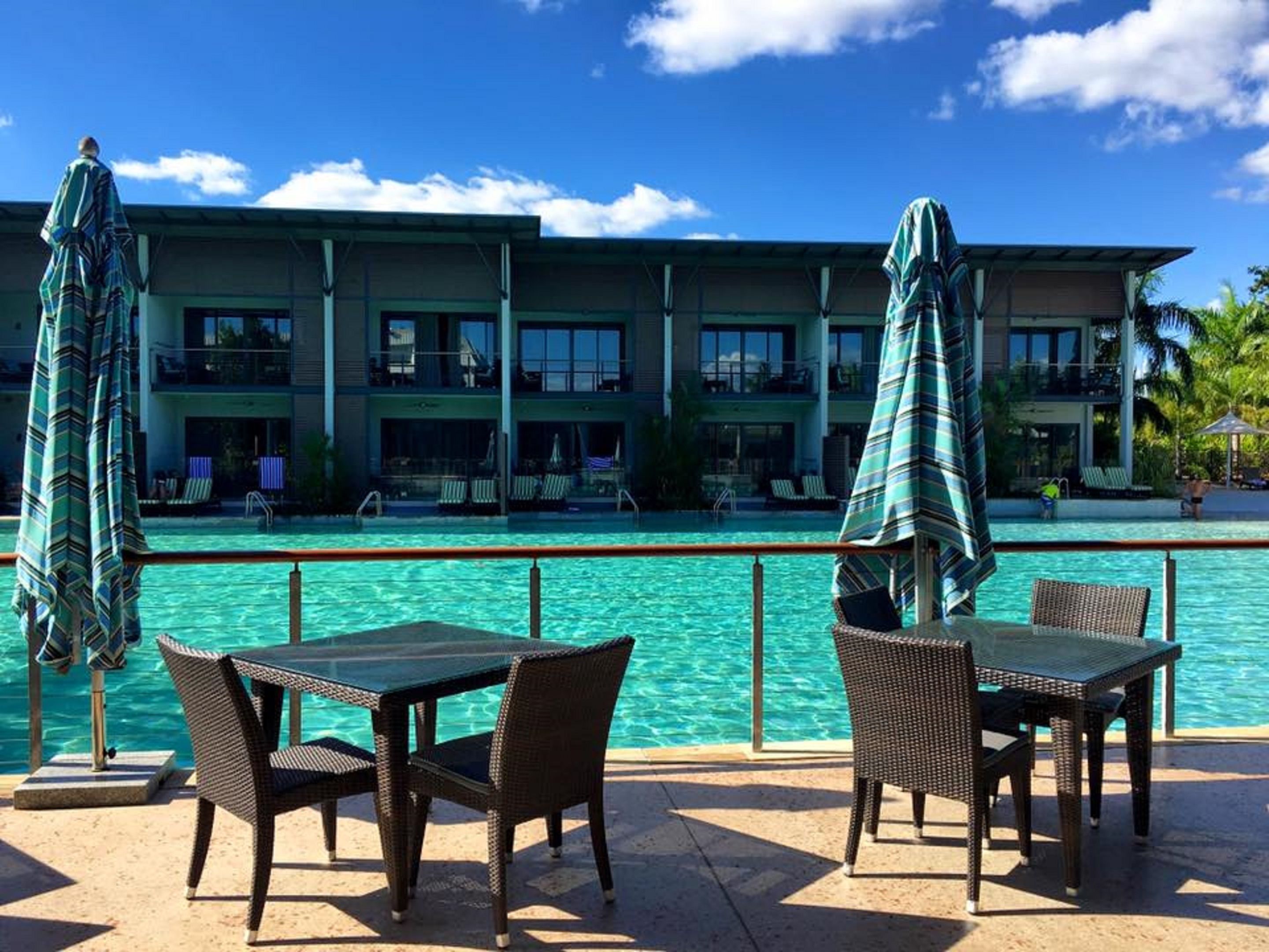 Un espace aménagé au bord de la piscine, avec des meubles en osier et des parasols, offrant une vue sur la piscine et un bâtiment doté de nombreux balcons.