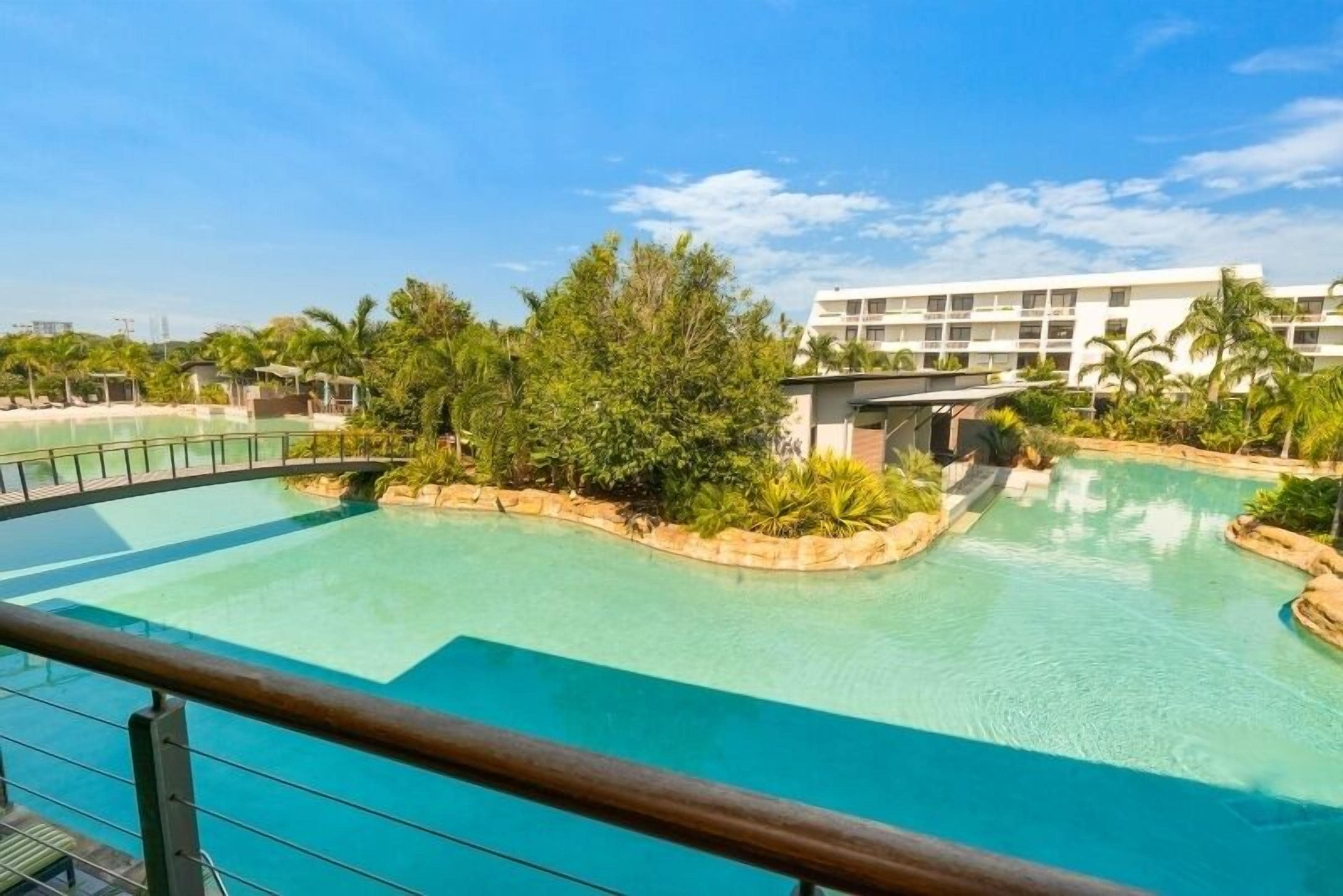 Ein Poolbereich eines Resorts mit klarem, blauem Wasser, umgeben von üppiger Vegetation und einem weißen Gebäude im Hintergrund.